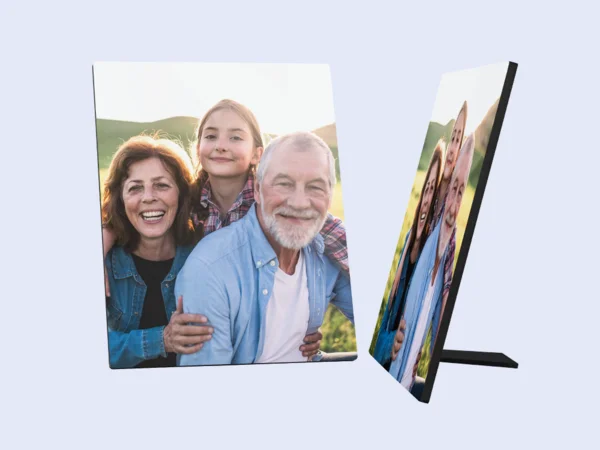 Ein Fotoaufsteller zeigt ein fröhliches Familienbild. Eine lächelnde ältere Frau mit braunen Haaren steht links. Hinter ihr steht ein junges Mädchen mit blonden Haaren in einem karierten Hemd und lächelt ebenfalls. Rechts von ihnen steht ein älterer Mann mit weißen Haaren und Bart, der auch fröhlich in die Kamera schaut und seine Arme um die beiden anderen gelegt hat.